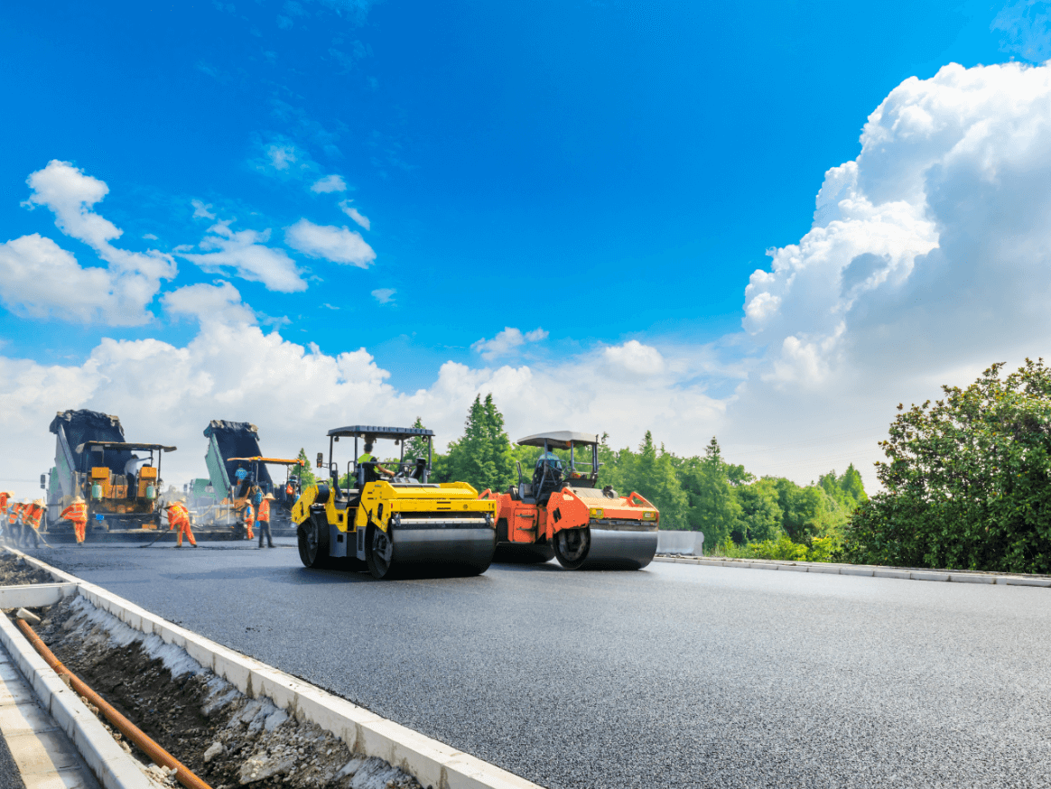 晴天の下で舗装工事を行う重機と作業員たちの現場風景
